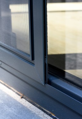 Otwarta moskitiera przesuwna na drzwi balkonowe. Skrzydło z siatką wykonaną z włókna szklanego oraz uszczelkami szczotkowymi które gwarantują szczelność pomiędzy ramą moskitiery drzwiowej a ramą okienną poruszające się po szynie jezdnej (prowadnicy).